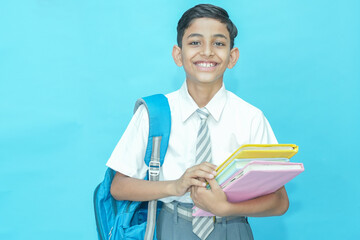 Happy Indian student schoolboy wearing uniform holding notebooks and bag standing isolated over...