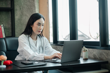 Female doctor in white coat using laptop in a well-lit office, focused on work.