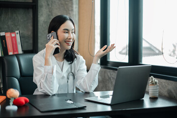 A cheerful female doctor in a white coat talks on her smartphone in a modern office setting with a...