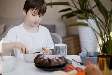Young boy having breakfast at home, eating cake at table with mugs and bowls, cozy morning