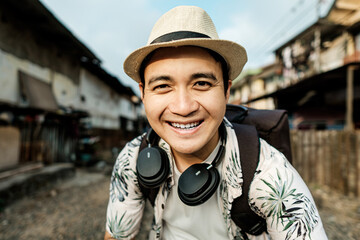 A cheerful traveler wearing a straw hat and headphones smiles brightly in a rural alley, ready to...