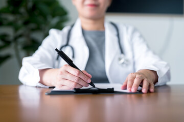 Smiling female doctor in lab coat with stethoscope writing on clipboard at desk, modern healthcare services, professional consultation, medical planning, and patient information recording.