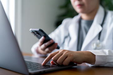 Doctor using smartphone and laptop together in clinical office. Modern digital healthcare communication, telemedicine, multitasking medical technology and efficient health data management.