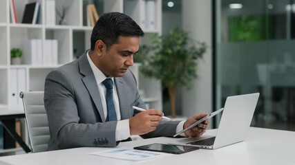 A focused indian businessman in a suit works diligently on his laptop and tablet in a modern office setting - Powered by Adobe