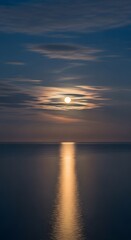 Fototapeta premium A serene night seascape featuring a full moon behind thin clouds, its brilliant golden light creating a shimmering reflection path on the calm, tranquil surface of the dark ocean