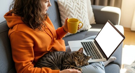 A young woman in an orange hoodie sits on a grey sofa, holding a yellow mug and using a laptop with a blank screen, while a tabby cat rests on her lap.
