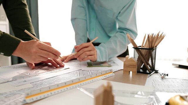 Smart architect and safety engineer discussion about building blueprint on table with architectural document, measurement tools and wooden block scatter around at modern office. Closeup. Delineation.