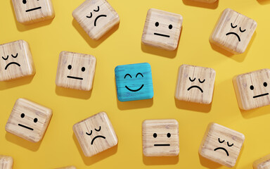 Blue wooden block with a smiling face stands out among sad and neutral face blocks on yellow background. Concept of positivity, mental health, uniqueness, or workplace morale.