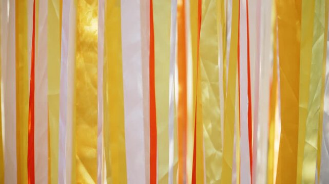 Background of moving multicolored vertical ribbons in orange, yellow and white. Bright festive multicolored silk stripes gently move in wind and in sunlight, abstract video.