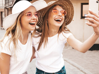 Two young smiling hipster blond women in summer white t-shirt. Female taking selfie self portrait...