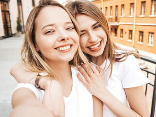 Two young smiling hipster blond women in summer white t-shirt. Female taking selfie self portrait...