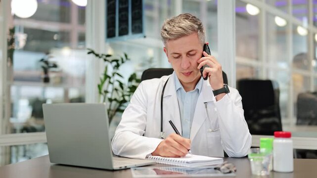 Focused medical professional takes important notes while on a phone call consultation at an office desk. Doctor is diligently working, providing medical advice and showing serious concentration.