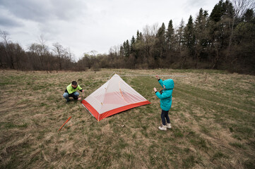 Camping adventure unfolds at a serene meadow in spring as a child assists an adult in setting up a vibrant tent under a cloudy sky