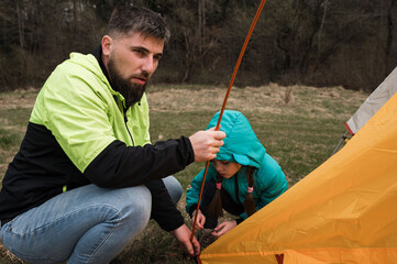 Family bonding while setting up a colorful tent in a serene meadow surrounded by trees during a...