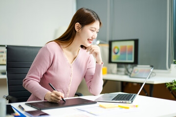 Asian designer using digital pen and laptop in a creative office, showcasing professional work,...