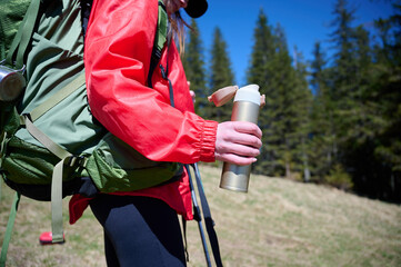 Cropped view of female hiker in red raincoat holding water bottle while trekking in mountain...