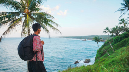 Solo hiker standing near grassy cliff overlooking turquoise sea and palm fringed coastline. Great...