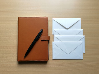 A brown leather notebook with a pen lies next to a stack of white envelopes