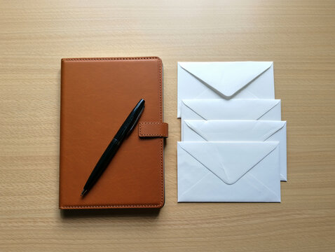 A brown leather notebook with a pen lies next to a stack of white envelopes