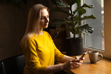 a woman stands near the window with her phone and morning coffee.