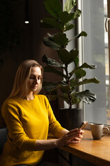a young woman gazes out the window with a coffee and a phone.