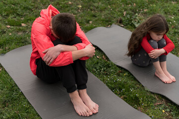 Two young individuals sit apart on yoga mats in a grassy park, lost in their thoughts as a gentle...