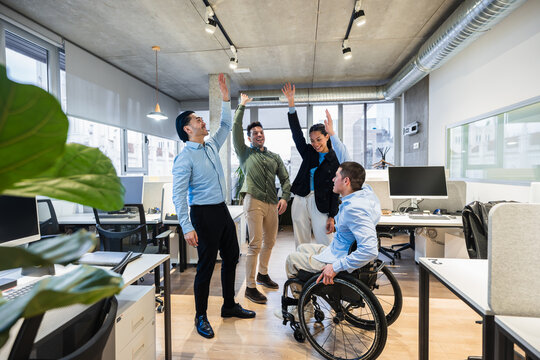Coworkers celebrating success, raising hands in modern office, showing inclusion and teamwork with disabled colleague in wheelchair - Powered by Adobe