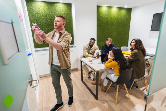 Young man writes on a glass board, leading a brainstorming session with a diverse team of colleagues in a modern coworking space - Powered by Adobe