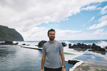 Man standing by natural volcanic swimming pools on a rugged coastline with crashing waves and a...
