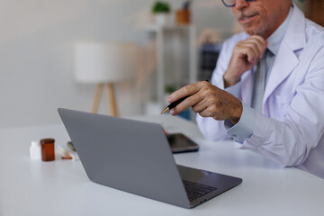 Senior middle aged male medical worker in modern clinic wearing white doctor's coat using laptop writing health personal data, consulting remotely. Telemedicine healthcare concept.