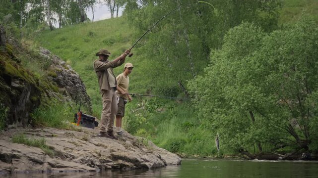 Wide shot of mature Asian dad standing on river bank with teenage son in summer, engaged in angling. Man catching wild fish on hook and pulling it out of water, while boy is clapping