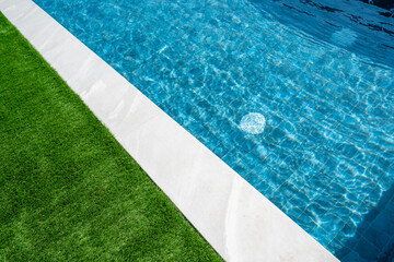 Obraz premium Swimming pool in the garden with white tiles around and green grass and clear transparent turquoise blue water. Real estate, property management and pool maintenance