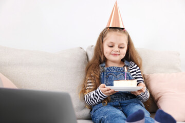 Birthday celebration. Little girl with piece of cake having video call via laptop on sofa at home