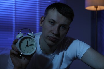 Frustrated man with alarm clock suffering from insomnia indoors
