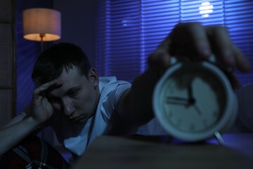 Frustrated man suffering from insomnia and turning off alarm clock indoors, selective focus