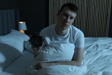 Frustrated man with alarm clock suffering from insomnia on bed indoors