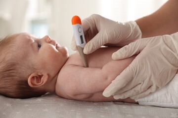Pediatrician examining baby with thermometer at table in clinic, closeup