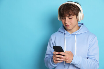 Cute teenage boy in headphones with smartphone on light blue background. Space for text