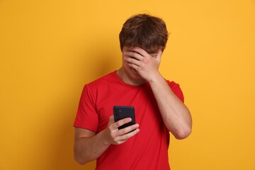 Teenage boy with smartphone covering face with hand on yellow background