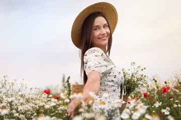 Smiling woman in hat with bag touching chamomile flowers in wildflower meadow