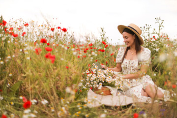 Smiling woman in hat with book and basket of chamomile flowers in wildflower meadow