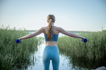 Young woman lifting dumbbells during outdoor workout by lake in summer