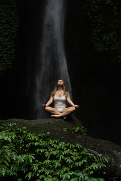 Young woman meditating in front of majestic waterfall in lush jungle