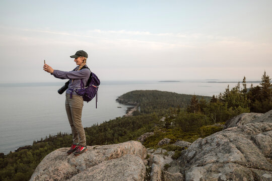 Female hiker on mountain top takes a picture with cell phone.