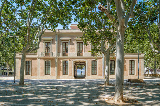 Neoclassical Building With Central Archway Inside Parc de Espanya Industrial Barcelona. Historic architecture, symmetrical facade, French doors, balconies, tree-lined plaza, summer shadows, park spain - Powered by Adobe