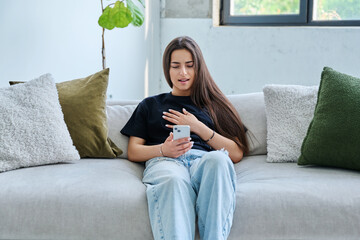 Young surprised woman sitting at home on couch using smartphone