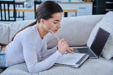 Young student girl study online, lying at home on couch with laptop, notebook