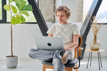 Handsome serious teenage boy sitting at home on the chair typing on laptop