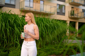 woman in casual attire holding beverage cup outdoors near modern apartment complex. smiling and looking away amidst lush greenery, conveying a relaxed urban lifestyle.