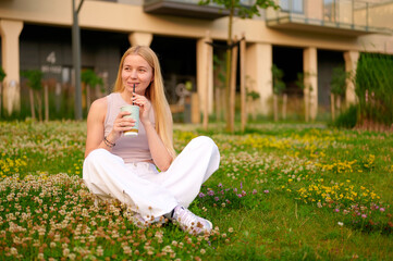 young woman enjoying cold drink while sitting on clover field. casual style, relaxed outdoor...